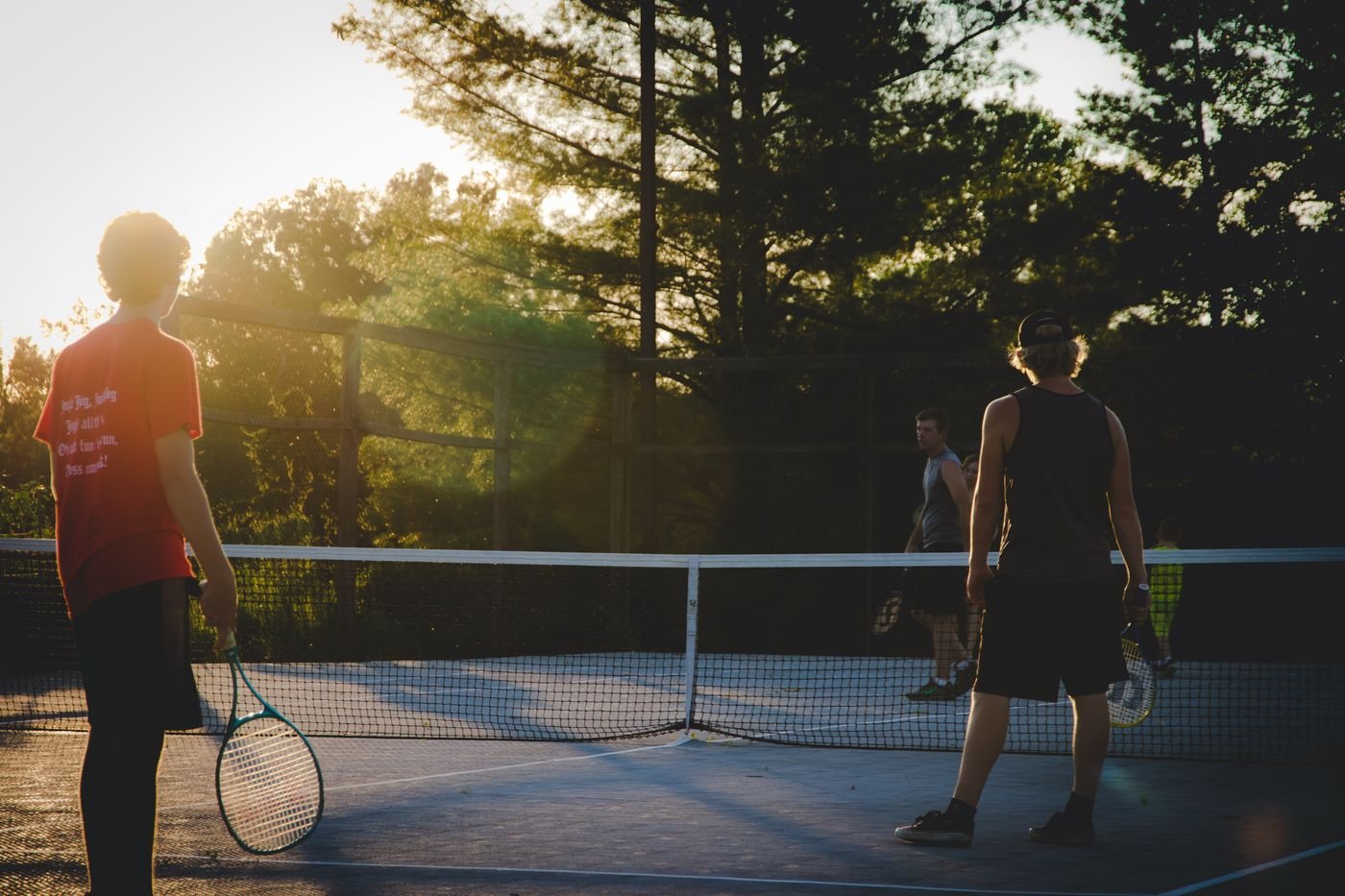 Players on a tennis court at sunset, seen from behind the net